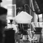 Black and white close-up of a barista pouring hot water into a Chemex coffee maker during a manual pour-over brewing process in a cozy café setting.