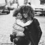 Black and white photo of a woman kneeling on a cobblestone street hugging a smiling young girl, capturing a warm family moment in an urban setting.