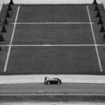 Black and white aerial photo of a lone person standing beside a car on a geometric road bordered by large grass sections and concrete lines, creating a minimalist urban composition.